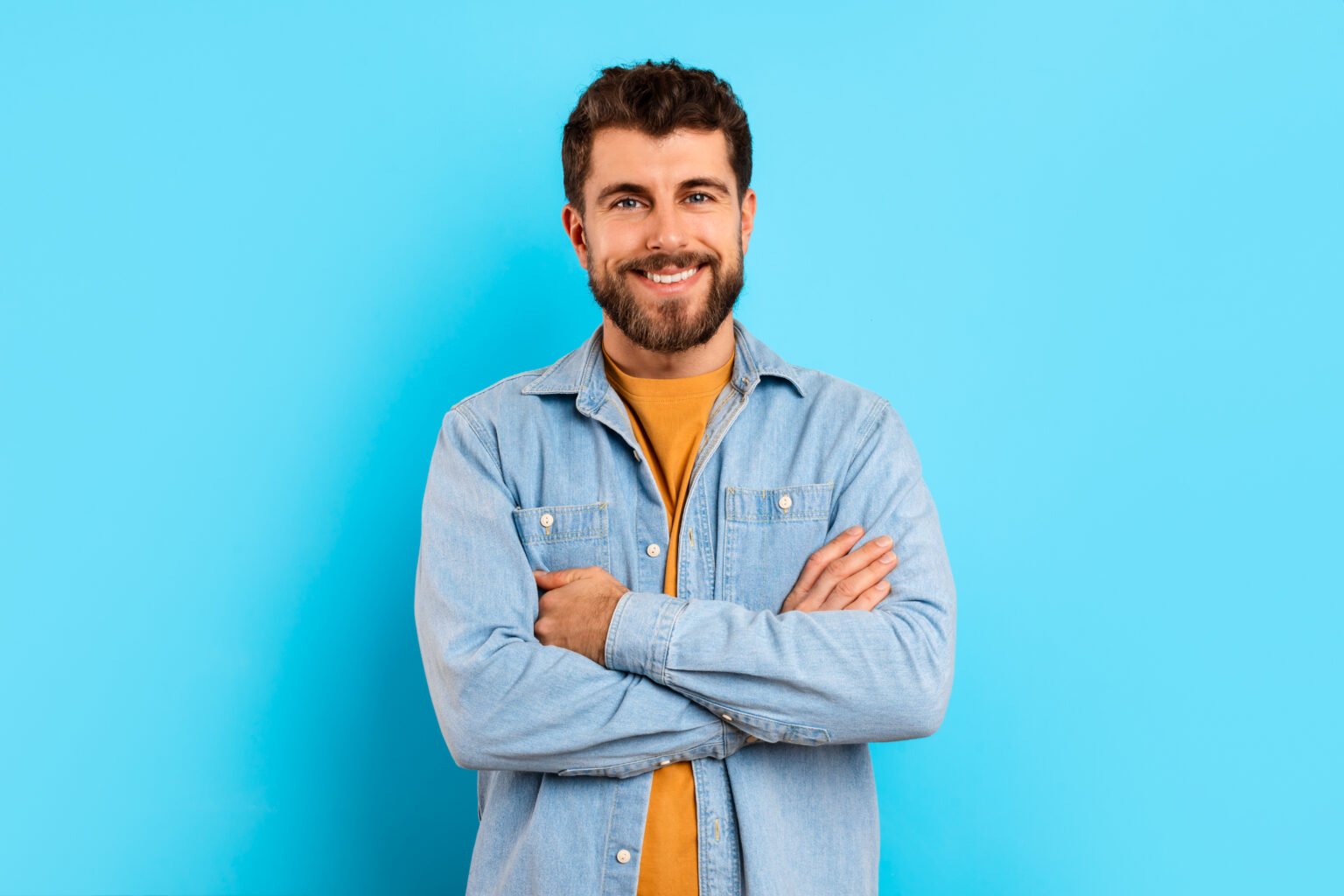 Portrait of cheerful confident young man in denim shirt, posing with arms folded smiling to camera, on blue studio backdrop. Banner with cheerful guy for advertisements and promotions
